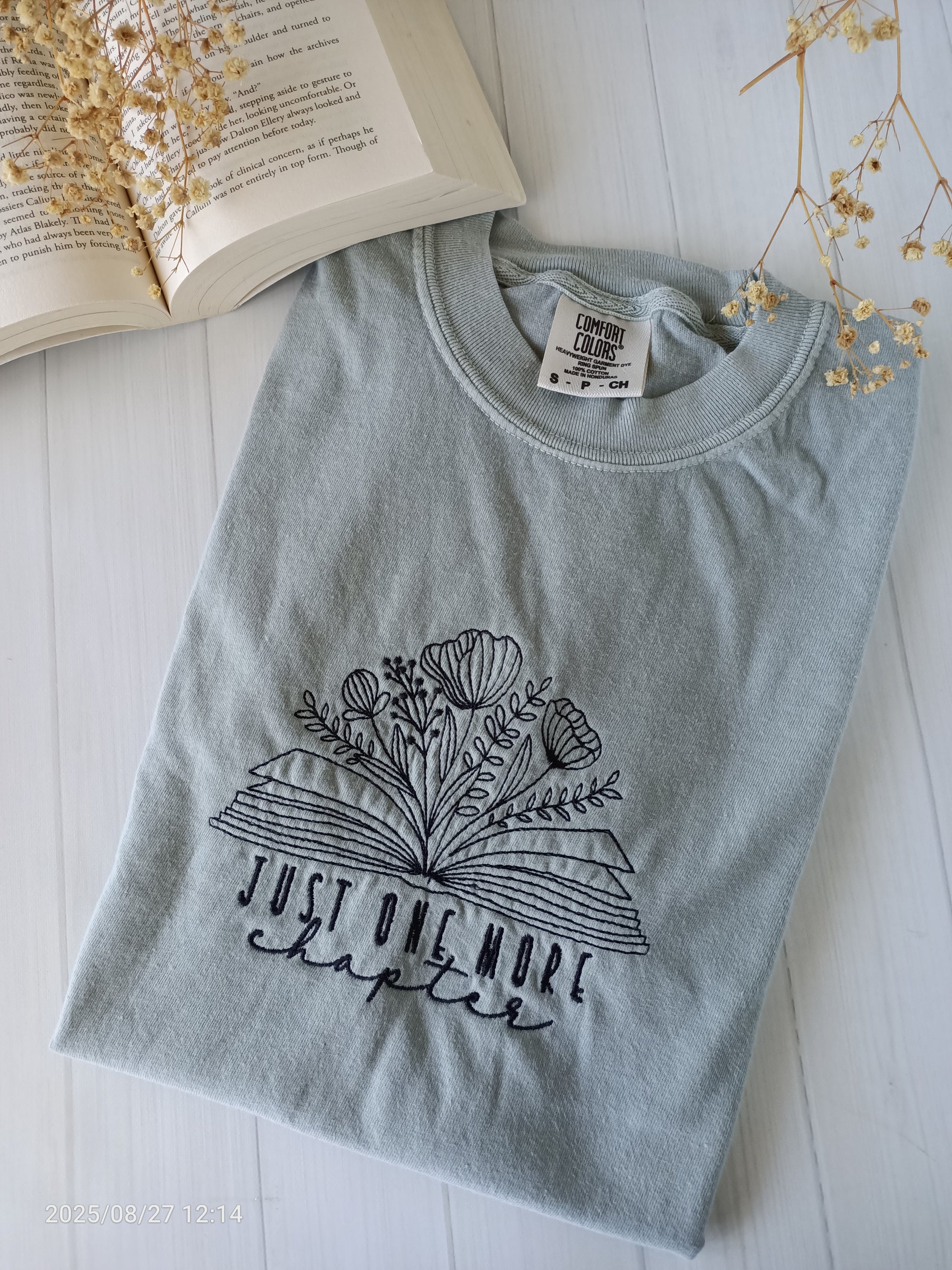 Sage green t-shirt with floral design and text 'Just One More Chapter' on a white surface with an open book and dried flowers.