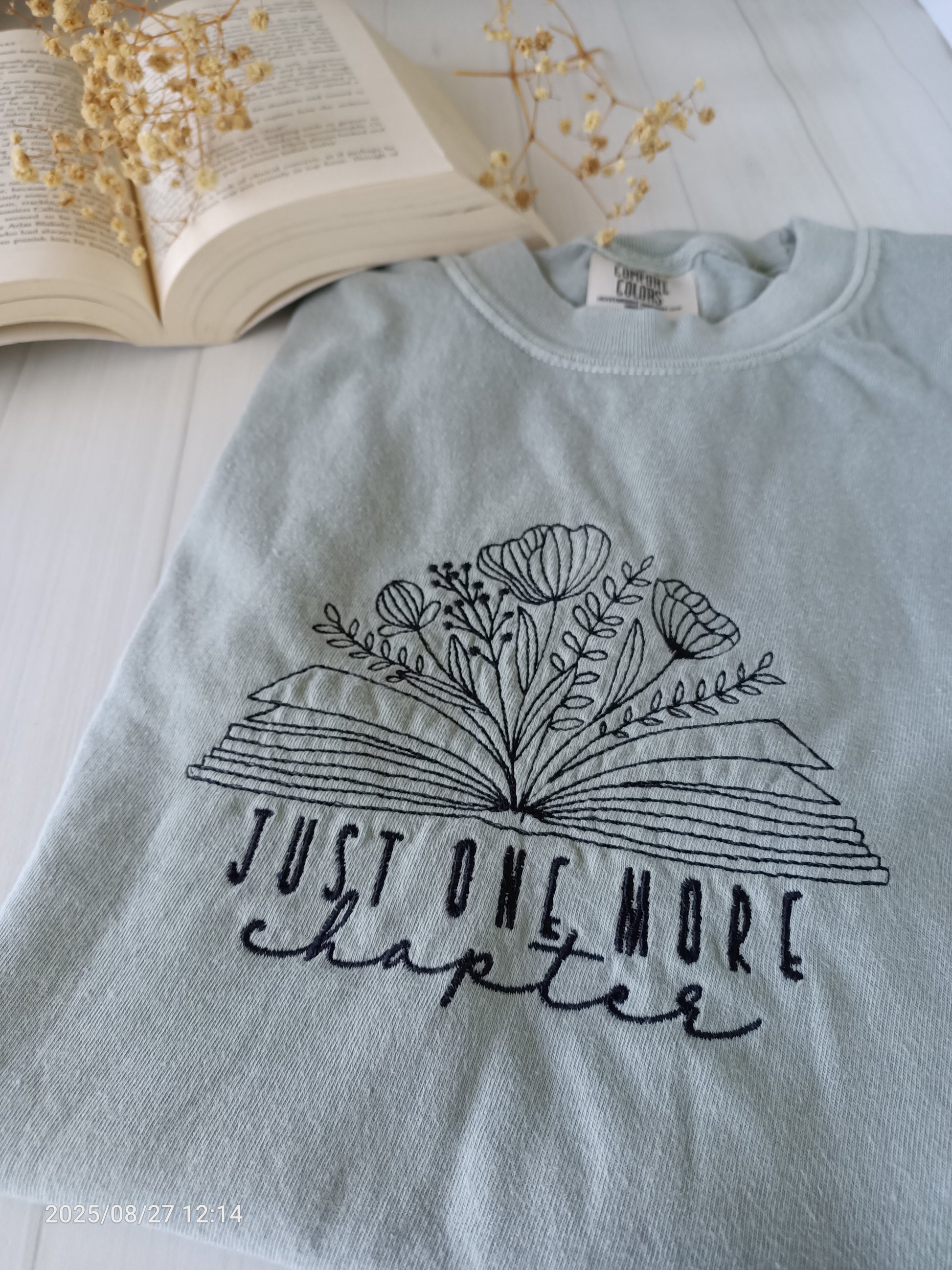 Sage green t-shirt with floral book design and 'Just One More Chapter' text, placed on a light surface with an open book and dried flowers.