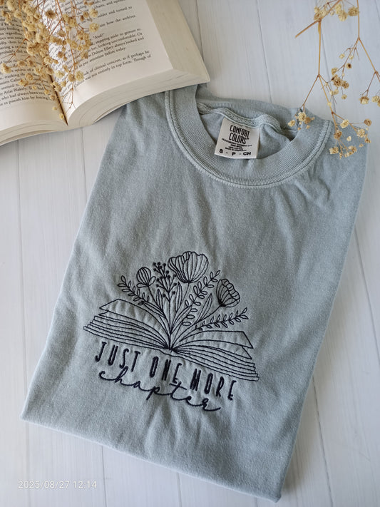 Sage green t-shirt with floral design and text 'Just One More Chapter' on a white surface with an open book and dried flowers.